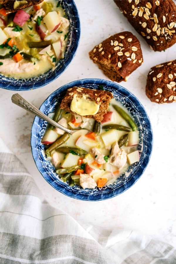 A bowl of vegetable soup with a piece of buttered bread on top. Another piece of bread and a second bowl of soup are in the background.