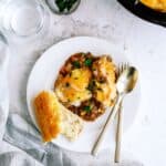 A plate of shepherd's pie topped with melted cheese, garnished with herbs, served alongside a slice of bread. A fork and spoon lie next to the food, with a glass of water and napkin nearby.