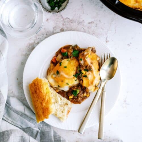 A plate of shepherd's pie topped with melted cheese, garnished with herbs, served alongside a slice of bread. A fork and spoon lie next to the food, with a glass of water and napkin nearby.