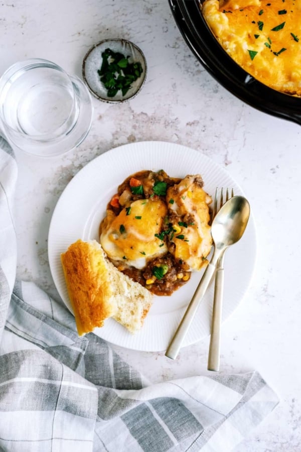 A plate of shepherd's pie topped with melted cheese, garnished with herbs, served alongside a slice of bread. A fork and spoon lie next to the food, with a glass of water and napkin nearby.