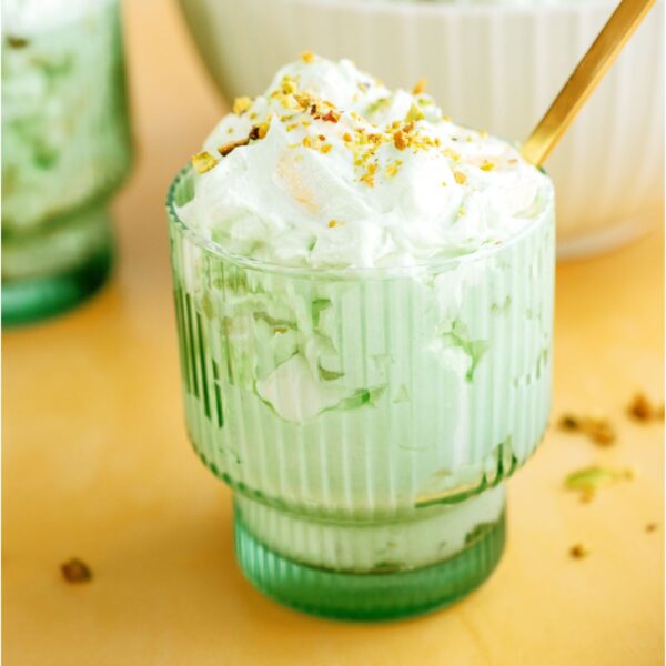 A green glass filled with a serving of Pistachio salad topped with sliced pistachios and a gold spoon. More 5 Minute Pistachio Salad in the background.