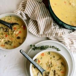 Two bowls of creamy soup with vegetables and mushrooms on a white table, next to a striped towel and a green pot filled with more soup.