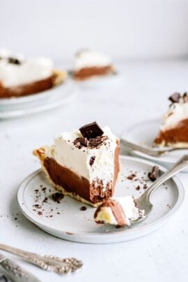 A slice of chocolate pie with whipped cream and chocolate shavings on a white plate, with a fork holding a small piece. Other slices are visible in the background.