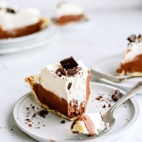 A slice of chocolate pie with whipped cream and chocolate shavings on a white plate, with a fork holding a small piece. Other slices are visible in the background.