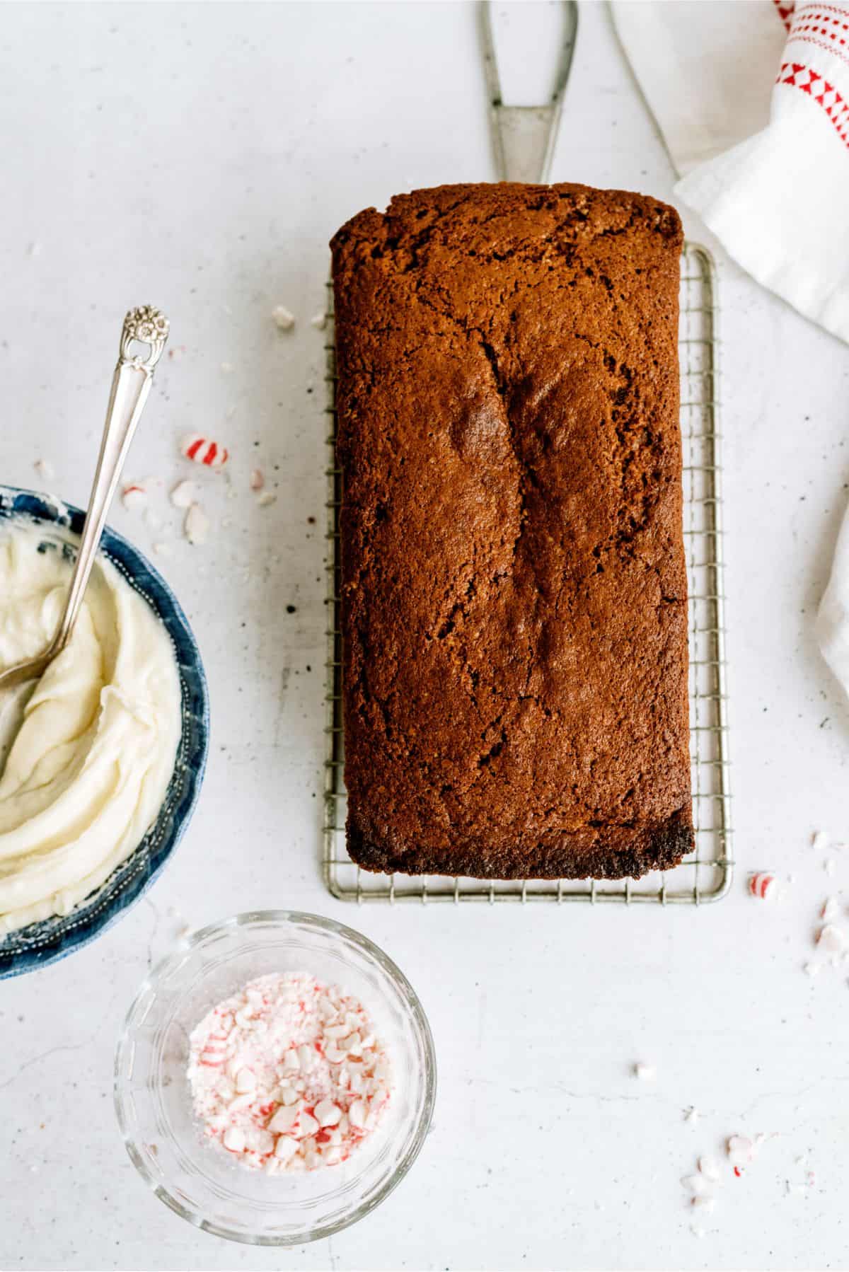 A loaf of cake on a cooling rack next to a bowl of frosting and a dish of crushed candy.