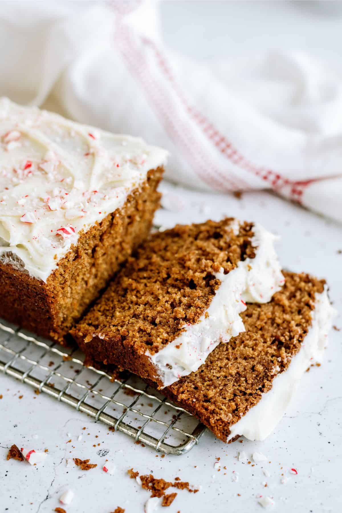 A sliced gingerbread loaf with white frosting on a cooling rack, sprinkled with crushed candy canes. A white cloth with red stripes is in the background.