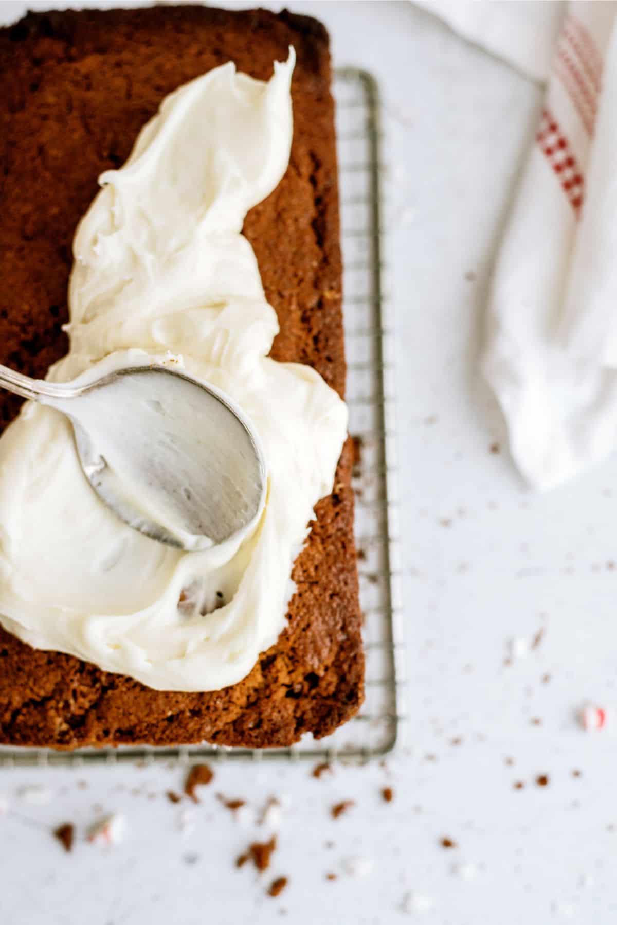 A loaf cake on a wire rack being frosted with white icing using a spoon.
