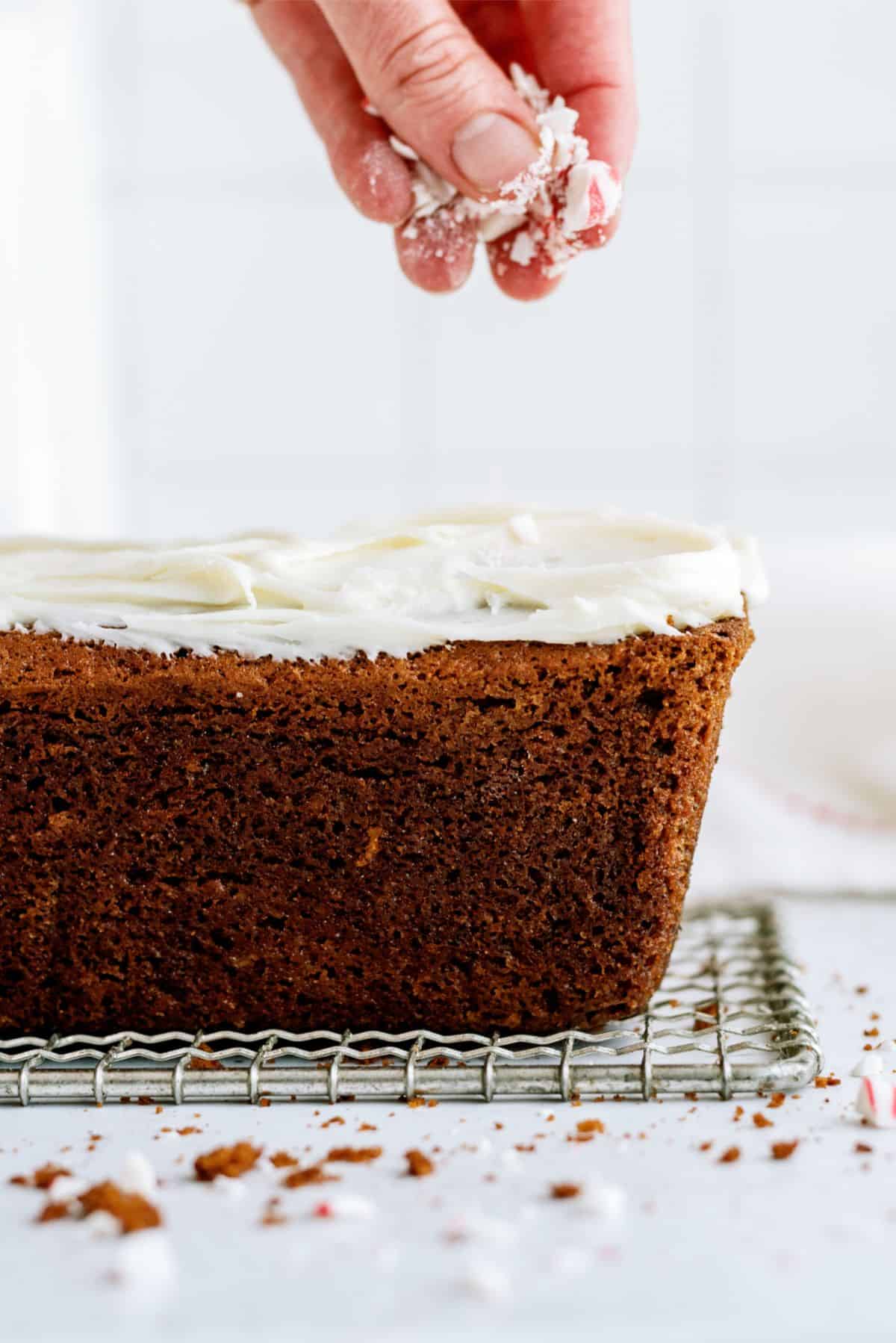 A hand sprinkles white frosting over a loaf cake on a cooling rack, with crumbs scattered on the surface below.