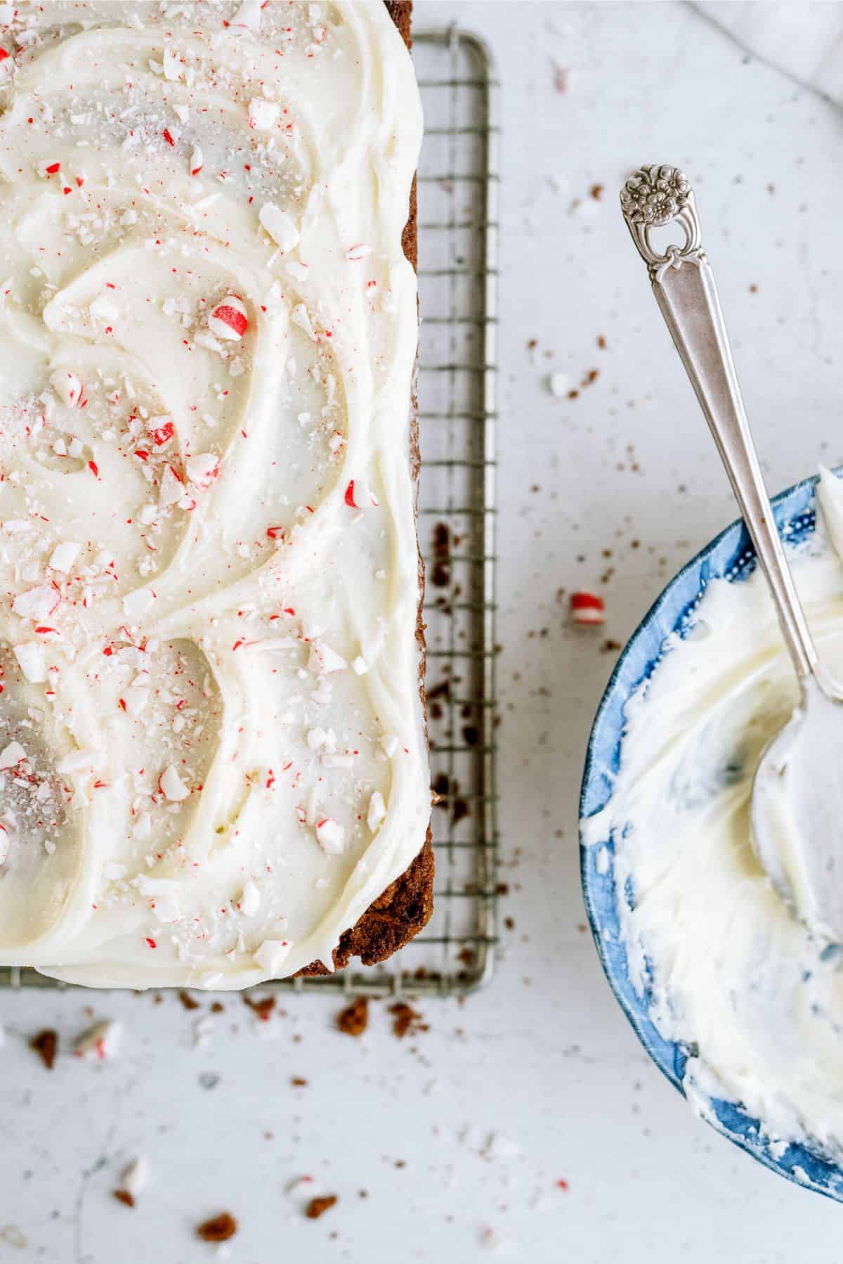 A frosted cake with crushed peppermint on a cooling rack, next to a bowl of partially used frosting.