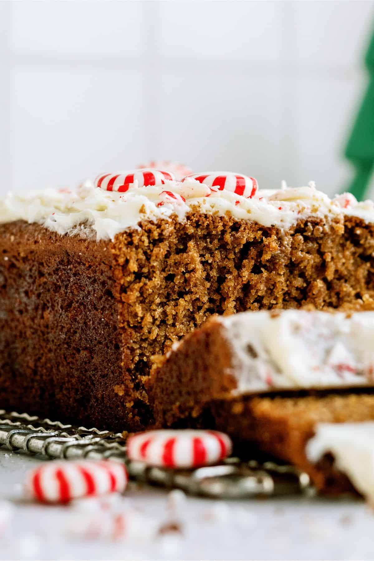 A slice of gingerbread loaf with white icing and peppermint candies on top, with one piece cut and laying in front.