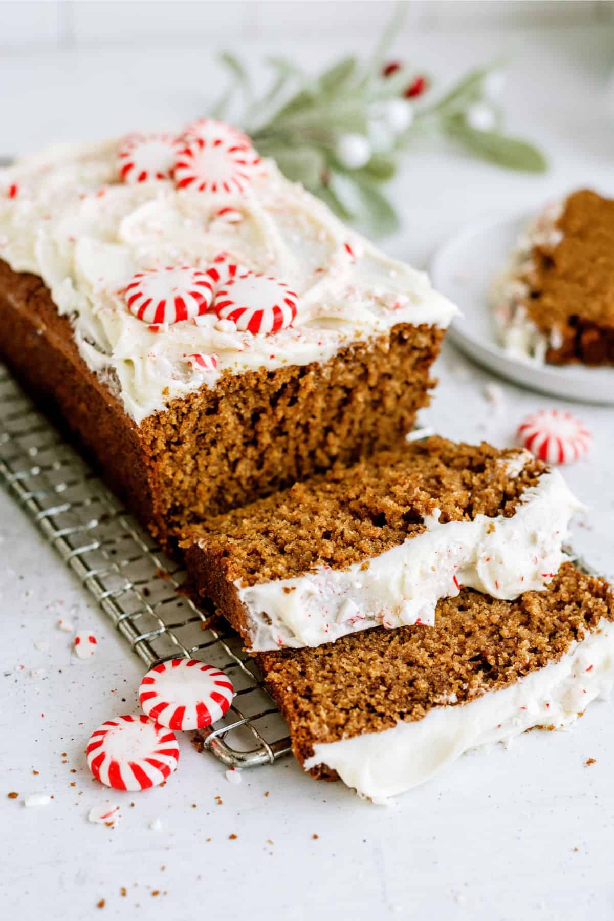 A loaf of gingerbread topped with white frosting and peppermint candies is sliced, with two slices in the foreground and a partially eaten slice on a plate in the background.