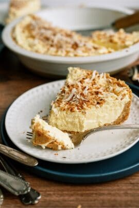 A plate with a slice of creamy, yellow pie topped with toasted coconut flakes. A fork with a bite of pie rests on the plate. The remaining pie is in the background.