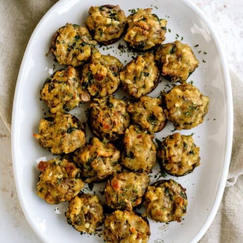 A white plate with several stuffed mushrooms topped with herbs, placed on a light surface beside a beige cloth napkin.