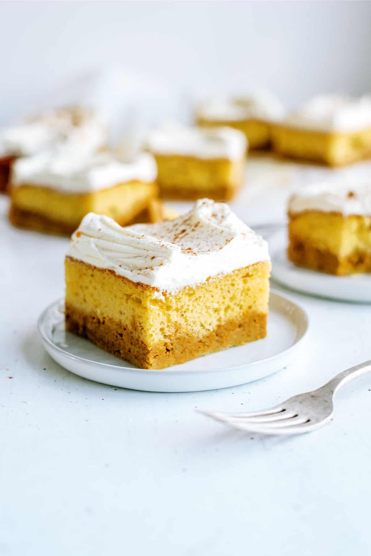 A small square pumpkin cheesecake bar with a graham cracker crust and whipped topping sits on a white plate. A fork is nearby, and similar bars are in the background.