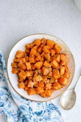 A white bowl filled with cubed and seasoned roasted sweet potatoes, placed on a light-colored surface with a metal spoon and a partially visible blue and white patterned cloth on the side.
