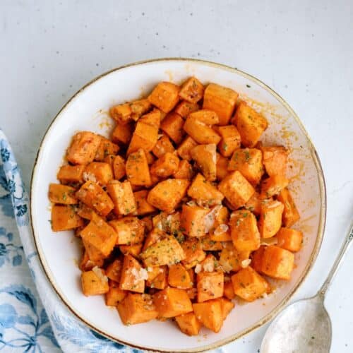A white bowl filled with cubed and seasoned roasted sweet potatoes, placed on a light-colored surface with a metal spoon and a partially visible blue and white patterned cloth on the side.