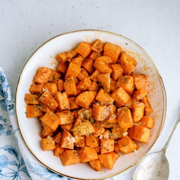 A white bowl filled with cubed and seasoned roasted sweet potatoes, placed on a light-colored surface with a metal spoon and a partially visible blue and white patterned cloth on the side.