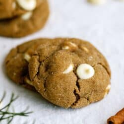 Two brown cookies with white chocolate chips on a white surface, with green sprigs partially visible.