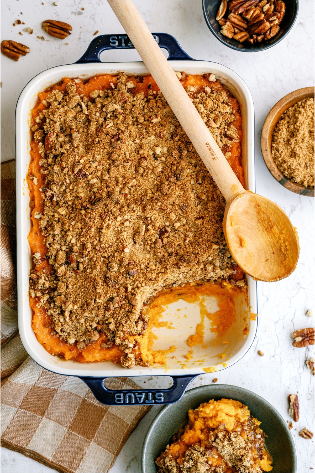 Sweet Potato Casserole in a casserole dish with a scoop missing and a wooden spoon on top of the casserole dish. A serving of Sweet Potato Casserole in a blue bowl with two small bowl of pecans and brown sugar in the background.