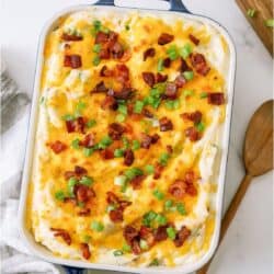 Baked casserole topped with melted cheese, chopped bacon, and green onions in a blue Staub dish on a white surface with a wooden spoon and cutting board nearby.