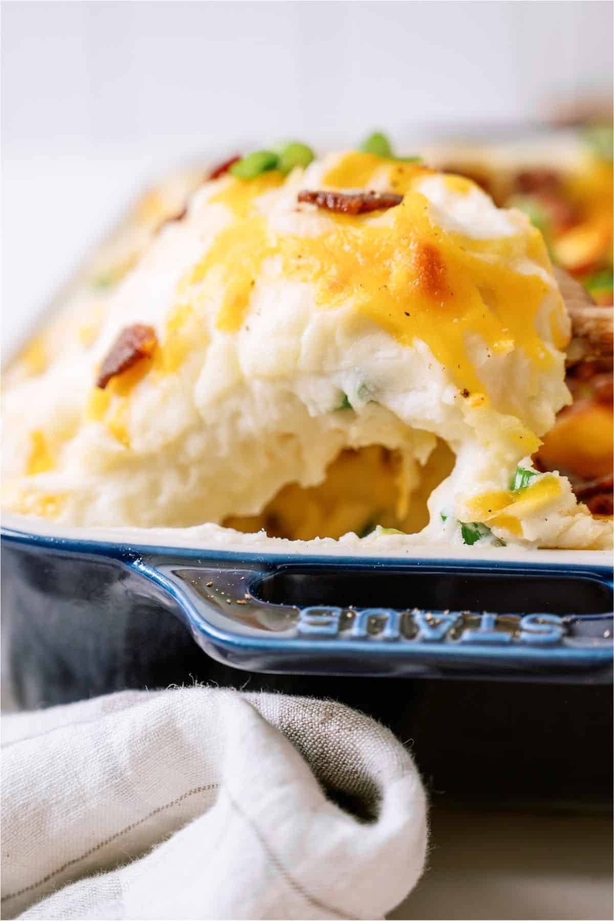 A wooden spoon lifting a scoop of Twice Baked Potato Casserole out of the baking dish.