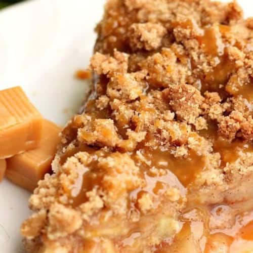 A slice of crumb-topped caramel apple pie on a white plate with caramel candies beside it.
