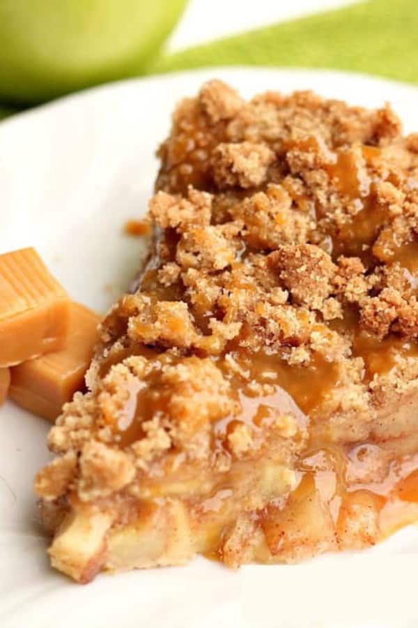 A slice of crumb-topped caramel apple pie on a white plate with caramel candies beside it.