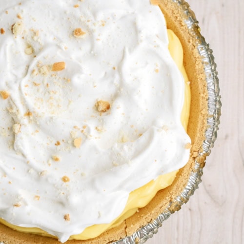 A close-up of a pie with a graham cracker crust, yellow custard filling, and a layer of whipped topping, sprinkled with crumbs, in a foil pie pan on a light wood surface.