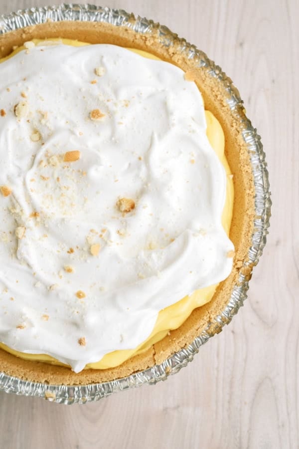 A close-up of a pie with a graham cracker crust, yellow custard filling, and a layer of whipped topping, sprinkled with crumbs, in a foil pie pan on a light wood surface.
