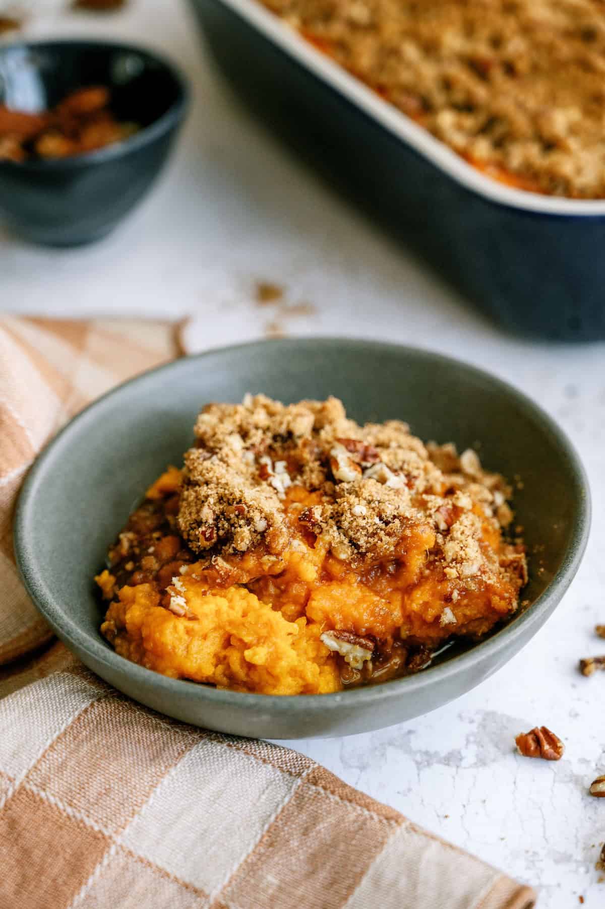 A bowl of sweet potato casserole topped with a brown sugar and pecan crumble, with a baking dish and a bowl of pecans in the background.