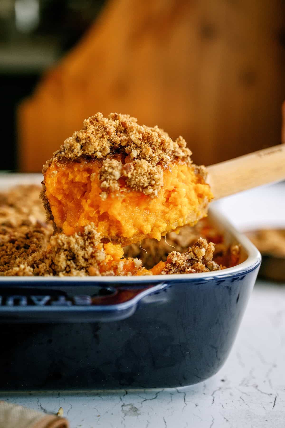 A close-up of a wooden spoon lifting a portion of sweet potato casserole with a brown sugar crumb topping from a blue baking dish.