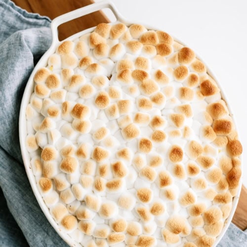 Oval casserole dish filled with sweet potato casserole topped with browned mini marshmallows, sitting on a blue cloth.