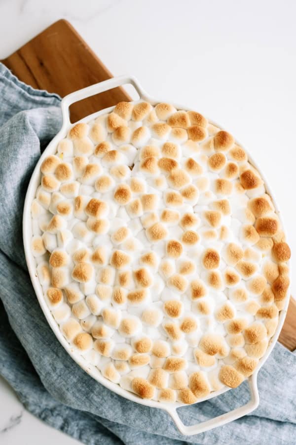 Oval casserole dish filled with sweet potato casserole topped with browned mini marshmallows, sitting on a blue cloth.