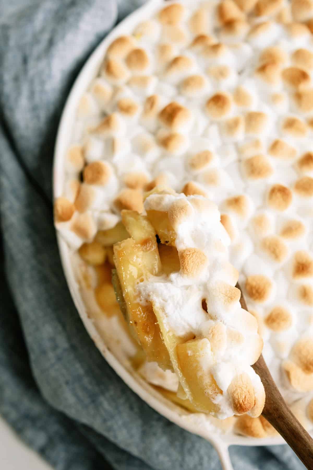 A close-up of a baked dessert topped with toasted mini marshmallows. A wooden spoon lifts out a serving, revealing slices of cooked apples underneath.