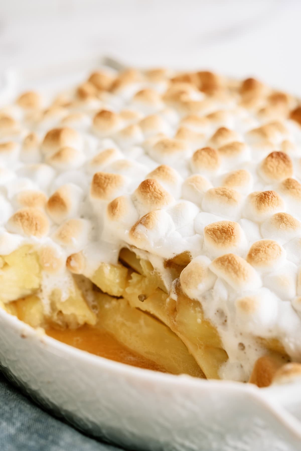 A close-up of a baked dessert featuring golden-brown toasted marshmallows on top of sliced apples in a white dish.