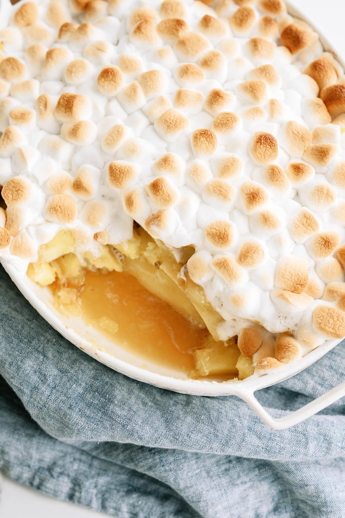 A baked dessert topped with toasted marshmallows, partially served to reveal a yellow filling and layers beneath, in a white dish on a blue cloth.