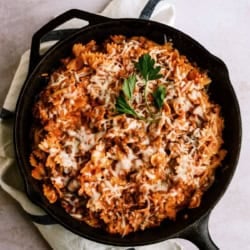 A cast iron skillet filled with bowtie pasta in a tomato meat sauce, topped with melted cheese and garnished with parsley, sits on a white towel.