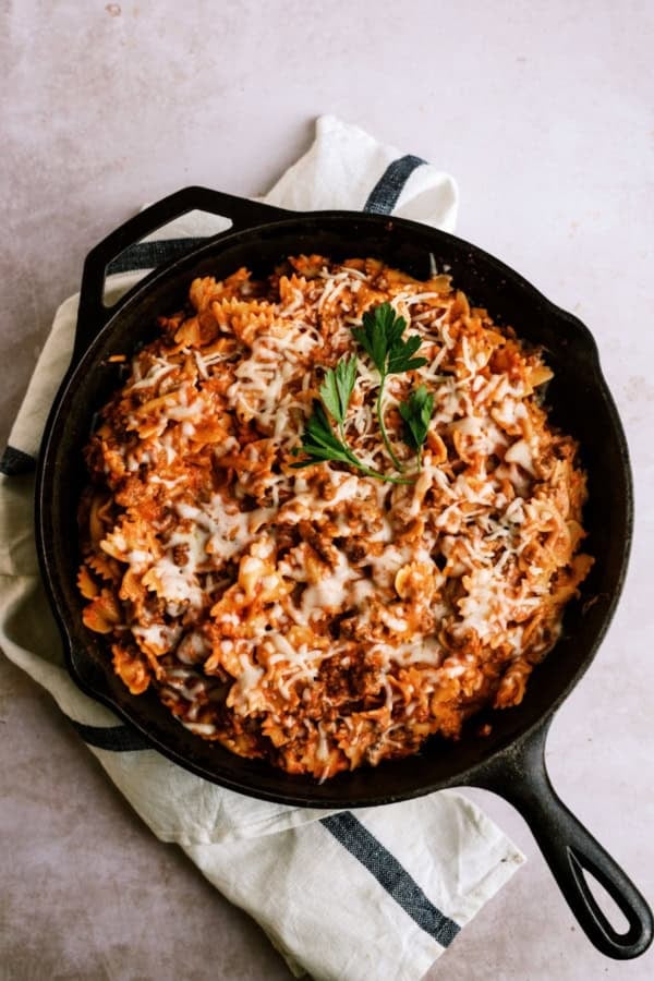 A cast iron skillet filled with bowtie pasta in a tomato meat sauce, topped with melted cheese and garnished with parsley, sits on a white towel.