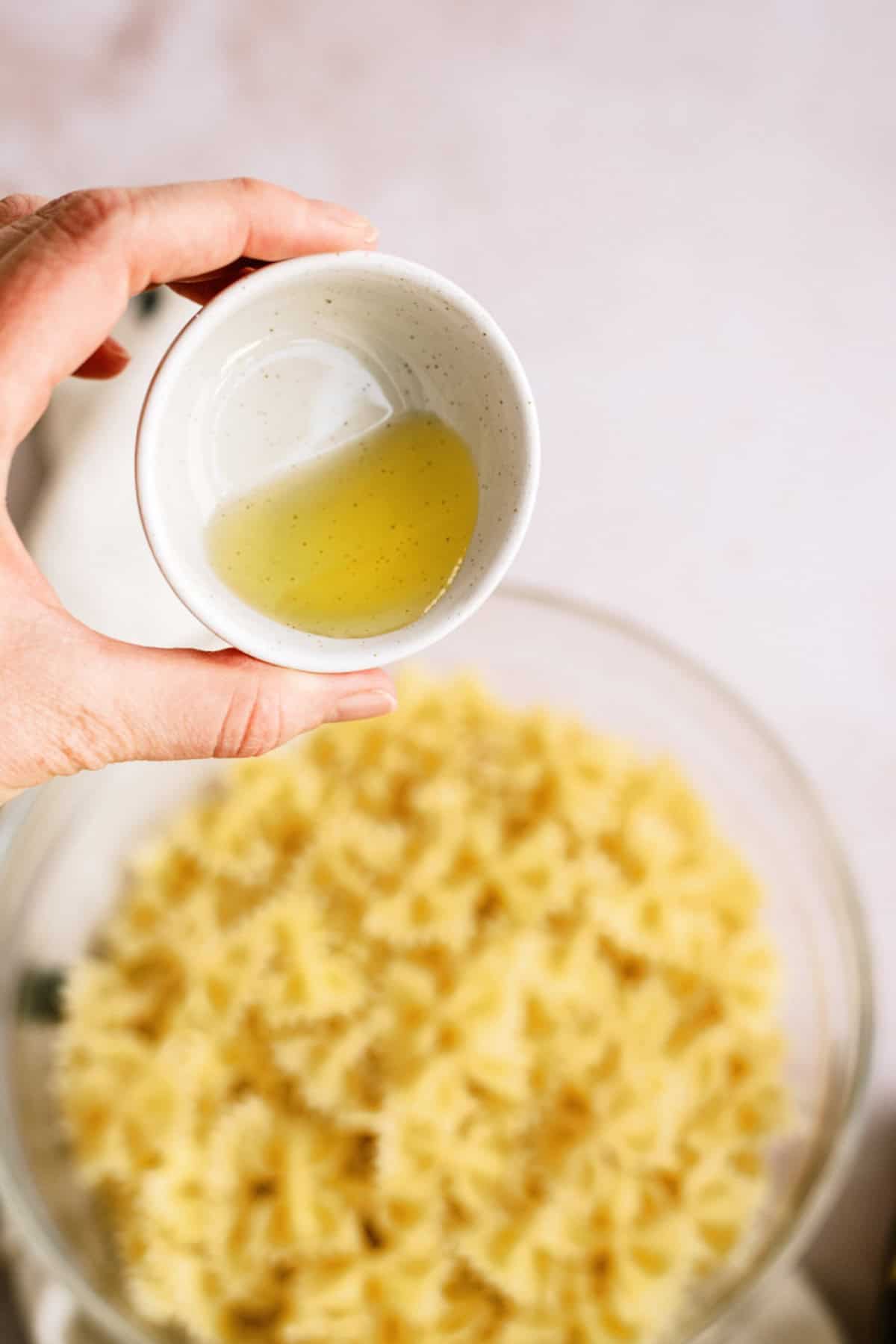 A hand holds a small bowl of olive oil above a glass bowl filled with cooked farfalle pasta.