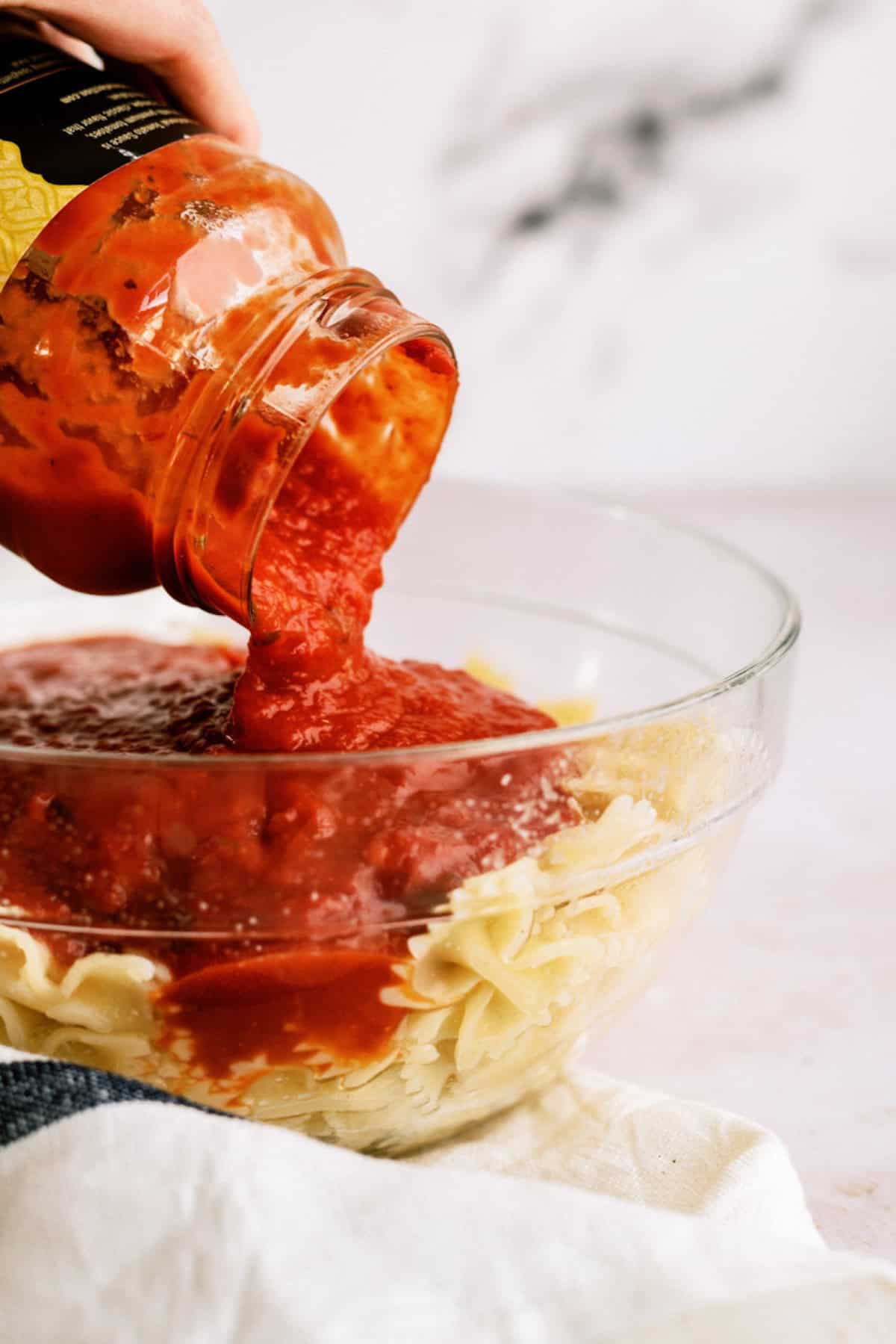 A hand pours tomato pasta sauce from a jar onto cooked rotini pasta in a glass bowl.