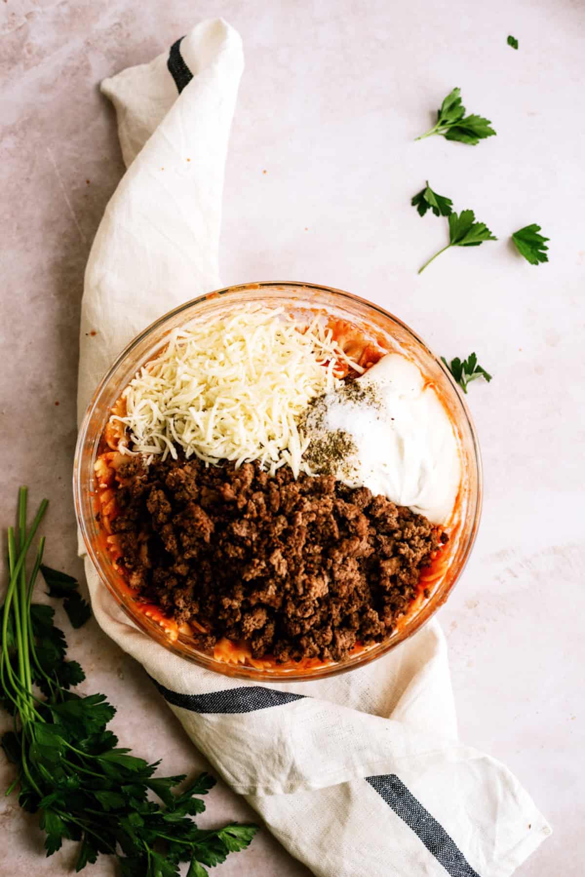 A glass bowl containing shredded cheese, ground cooked meat, sour cream, tomato sauce, and seasonings, with fresh parsley and a white kitchen towel nearby.