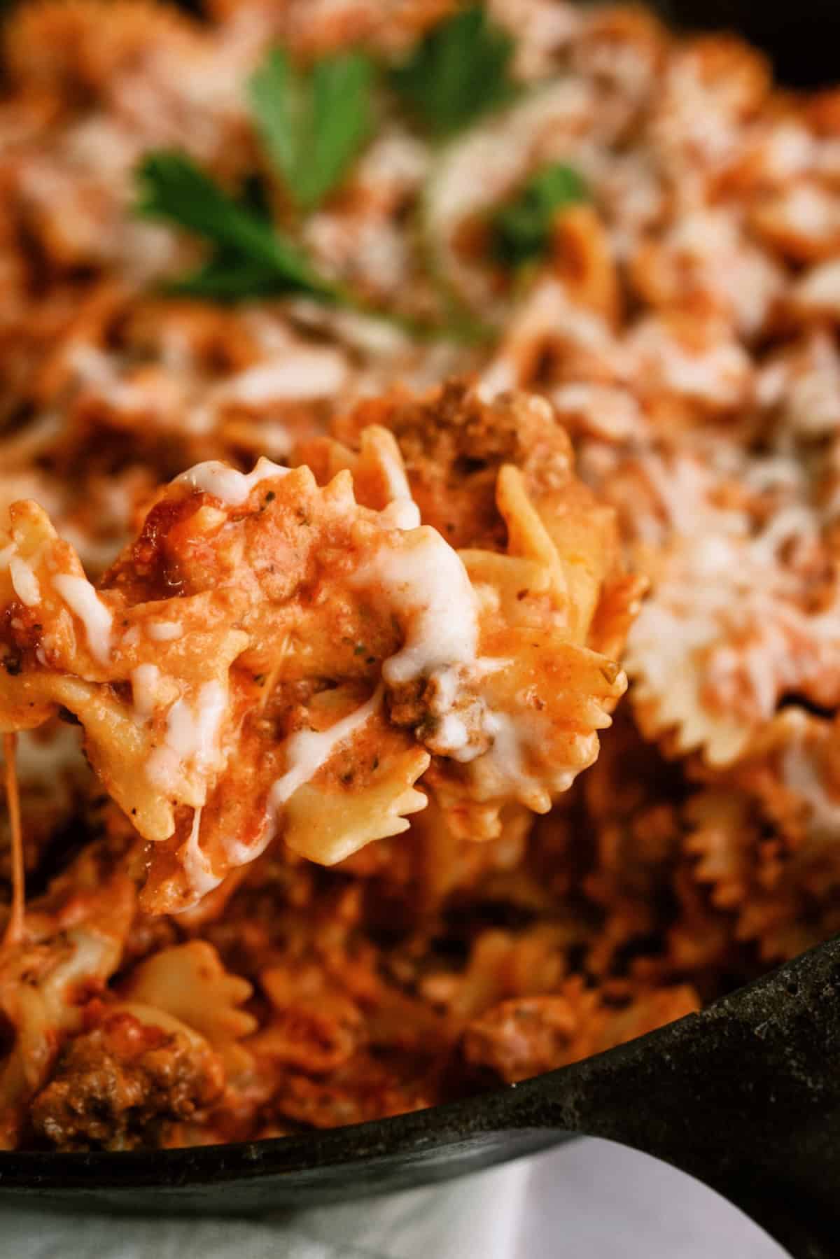 A close-up of bowtie pasta with ground meat, tomato sauce, and melted cheese in a skillet, garnished with fresh parsley.