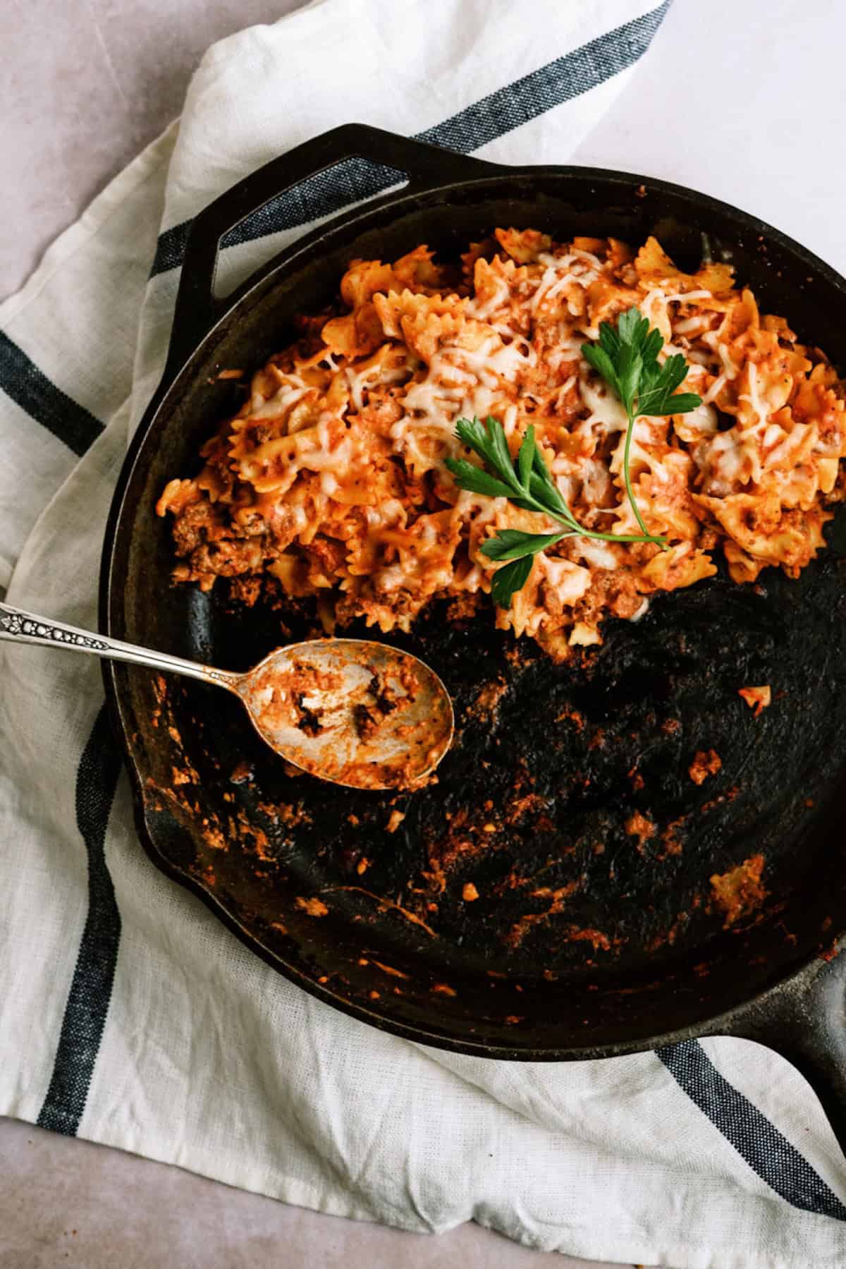 A cast iron skillet with baked pasta in tomato sauce, topped with melted cheese and parsley, sits on a cloth napkin with a serving spoon in the pan.