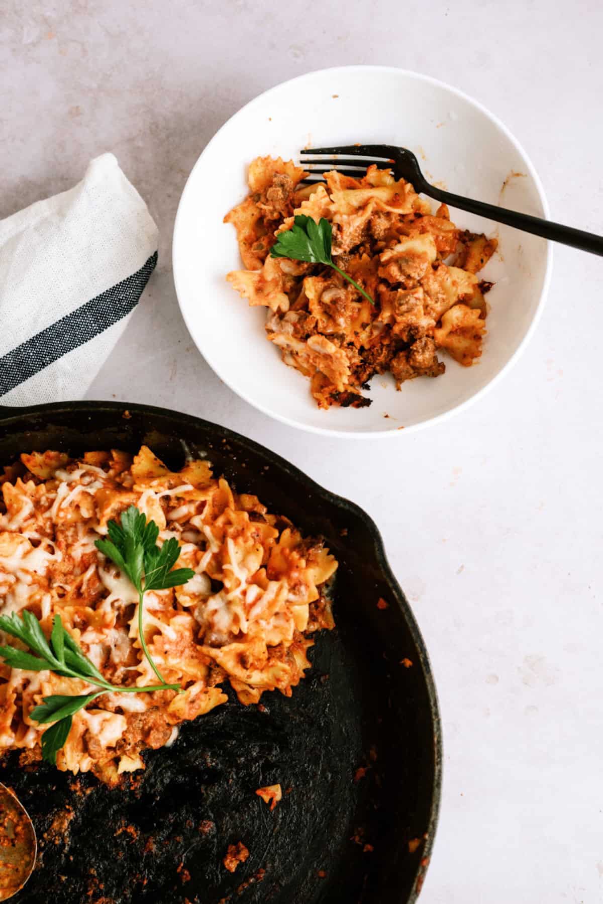 A serving of baked pasta with cheese and parsley in a white bowl next to a cast iron skillet containing the remaining pasta.