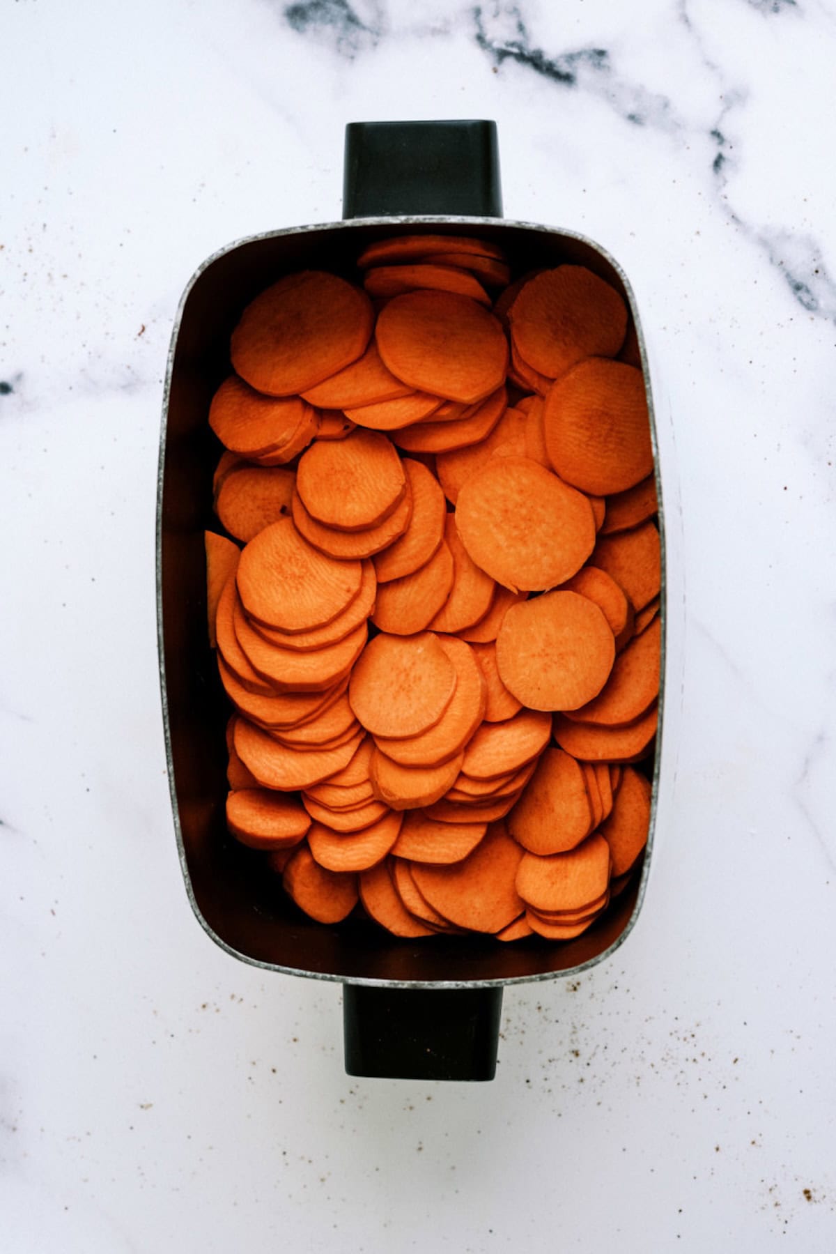 A rectangular black crockpot filled with thinly sliced raw carrots on a white marble surface.