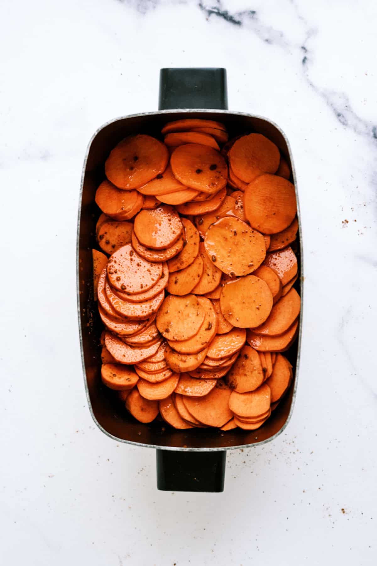 A rectangular baking dish filled with thinly sliced sweet potatoes seasoned with pepper, placed on a white marble surface.