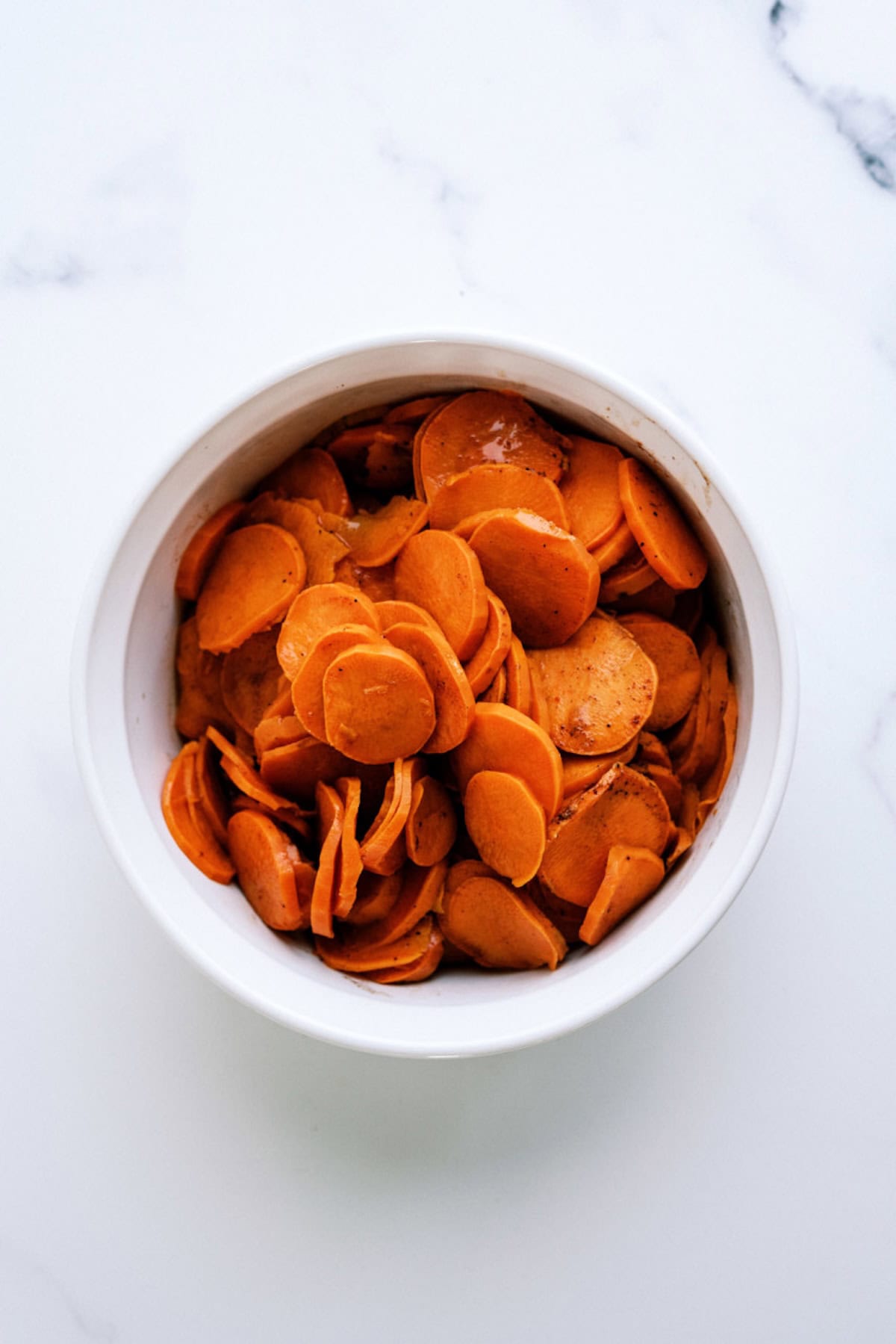 A white bowl filled with thinly sliced, seasoned sweet potatoes sits on a white marble surface.