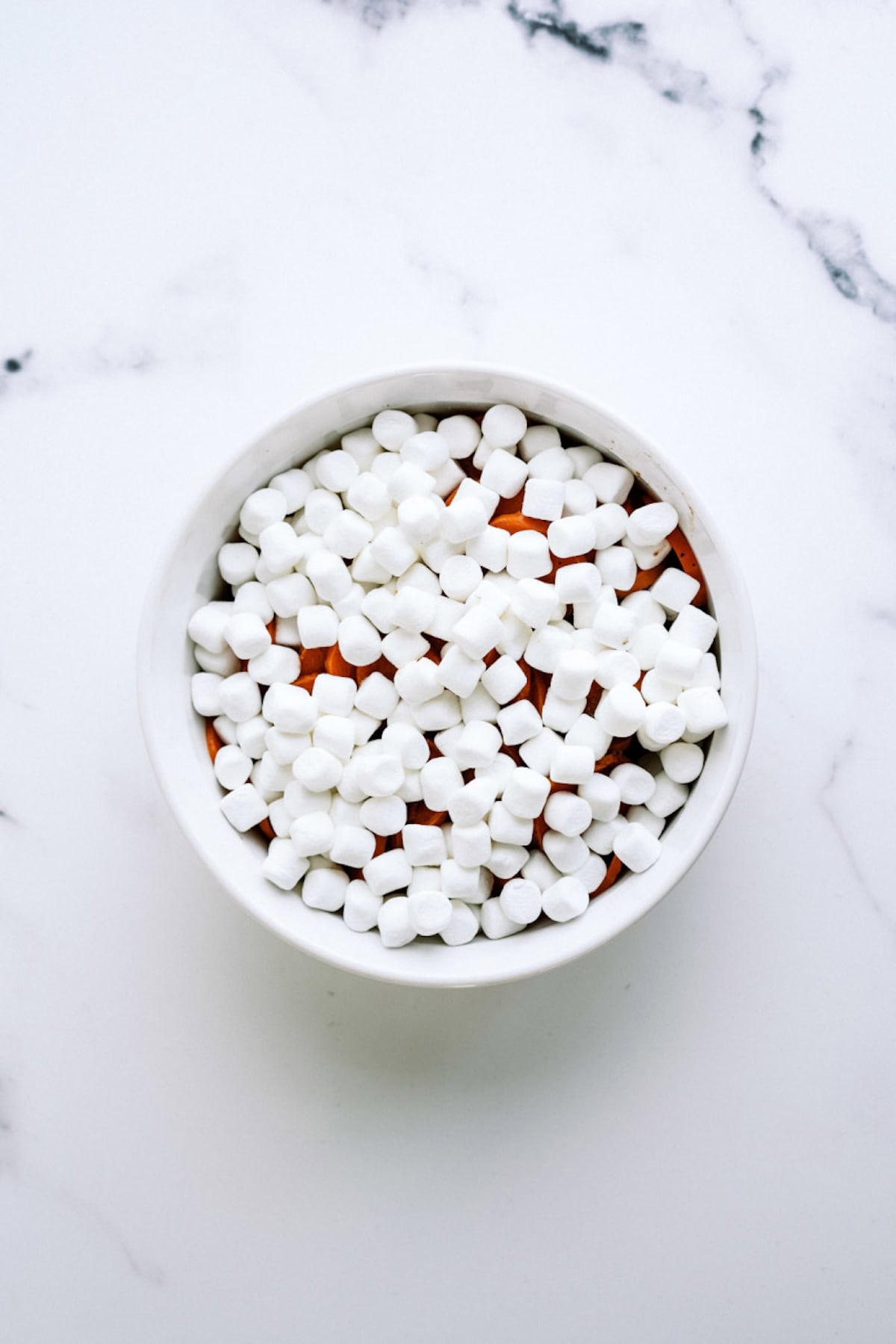 A white bowl filled with mini marshmallows sits on a white marble surface.