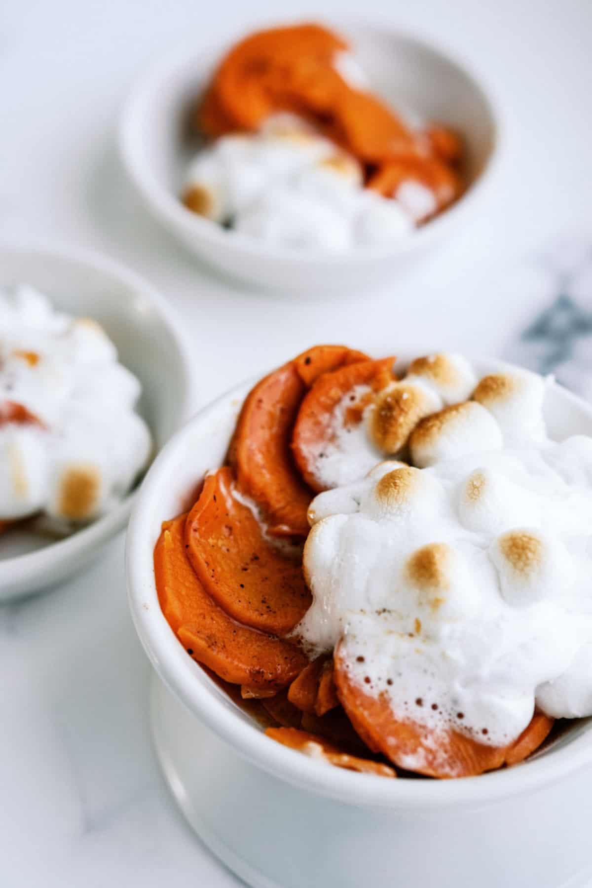 Three white bowls filled with sliced sweet potatoes topped with toasted marshmallows sit on a white surface.