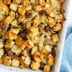 Golden brown bread stuffing with sausage and herbs baked in a white casserole dish, resting on a light blue cloth.
